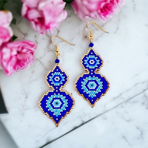 Puede incluir: Un par de pendientes de cuentas azules y doradas con un patrón geométrico. Los pendientes son de estilo colgante y presentan un diseño de ojo azul y blanco.