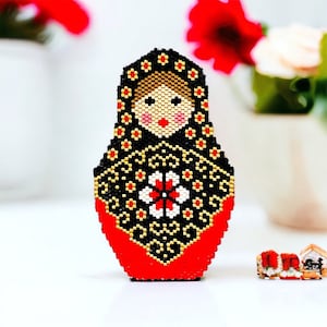 May include: A beaded Russian nesting doll, or matryoshka doll, with a black, gold, and red design. The doll has a red face with black eyes and a red flower on its chest.