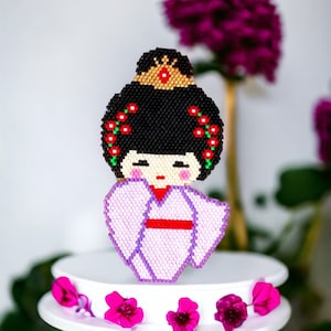 May include: A purple beaded doll with black hair, a red sash, and a gold crown. The doll is wearing a purple kimono with red flowers on the shoulders. The doll is sitting on a white cake stand with pink flowers.