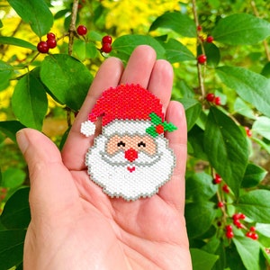 May include: A beaded Santa Claus ornament with a red hat, white beard, and green holly. The ornament is held in a hand against a background of green leaves and red berries.