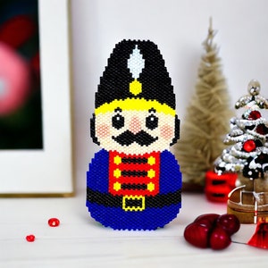 May include: A blue and red beaded nutcracker ornament with a black hat and a white plume. The nutcracker has a yellow face with black eyes, a black mustache, and a red mouth.