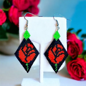 May include: A pair of beaded earrings with a red rose design. The earrings are diamond-shaped and have a green bead at the top. The earrings are hanging from silver hooks.