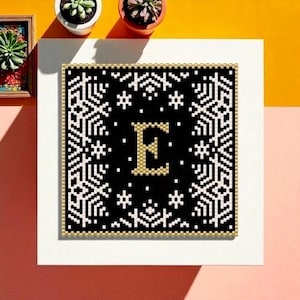 May include: A framed, square, black and gold beaded artwork featuring the letter "E" in gold. The artwork is surrounded by a border of white snowflakes and stars. The frame is white with a gold border.