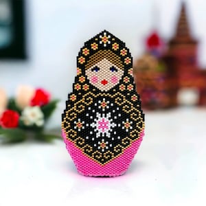 May include: A black and pink beaded Russian nesting doll, or matryoshka doll, with a gold floral pattern. The doll has a white and pink face with black eyes and a red mouth.