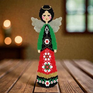 Peut inclure: Figurine d'ange perlée avec un halo noir, des cheveux noirs et des ailes grises. L'ange porte un haut vert et une jupe rouge avec des motifs floraux blancs. La figurine est décorée de perles noires, blanches et dorées et repose sur une surface en bois.
