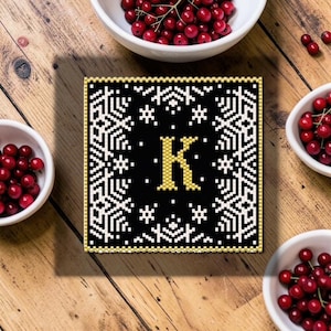 May include: A square black and white decorative tile with the letter "K" in gold. The tile has a snowflake design around the edges. The tile is surrounded by small white bowls filled with red berries on a wooden surface.