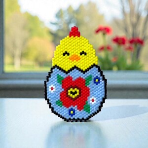 Peut inclure: Décoration de Pâques perlée représentant un poussin jaune sortant d'un œuf bleu orné d'une fleur rouge et de petites fleurs bleues et blanches. Le poussin a une crête rouge et un bec jaune. L'œuf est souligné en noir.