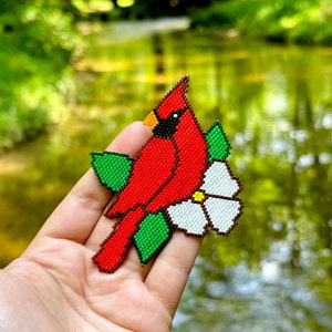 May include: A red cardinal bird with a white flower and green leaves, all made of beads. The beaded design is held in a person's hand.