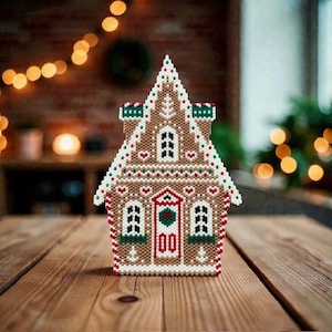 May include: A handcrafted gingerbread house decoration with a detailed, beaded design. The house features a brown base, white roof, and red, green, and white accents. The front door has a red frame and a green wreath. The windows are framed in white.
