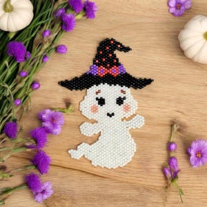 May include: A beaded Halloween ghost wearing a black witch hat with a purple bow. The ghost has rosy cheeks and a small smile. The image is set on a wooden surface with purple flowers and small white pumpkins.