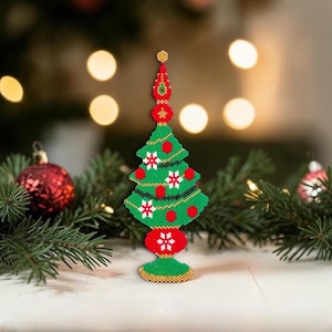 Può includere: Un ornamento per l'albero di Natale fatto di perline. L'albero verde è decorato con ornamenti rossi e dettagli di fiocchi di neve bianchi. La base e la parte superiore sono rosse con dettagli dorati. L'ornamento è su una superficie bianca con decorazioni natalizie sullo sfondo.