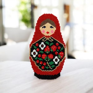 May include: A handcrafted, beaded Matryoshka doll. The doll features a red hood and a black apron adorned with red roses, white flowers, and green leaves. The doll's face has blue eyes, pink cheeks, and red lips. The base is black.