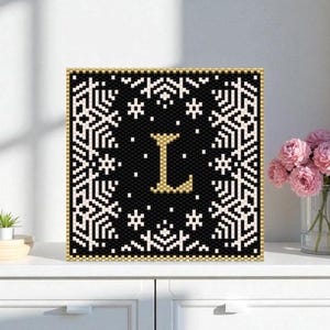 Op de afbeelding: Een vierkant, geborduurd kunstwerk met een zwarte achtergrond en een gouden letter 'L' in het midden. Het heeft witte sneeuwvlokpatronen en een gouden rand. Het stuk wordt getoond op een wit oppervlak, wat duidt op een decoratief item.