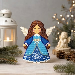 Könnte beinhalten: Ein Perlenengel-Ornament mit langem braunem Haar, weißen Flügeln und einem blauen Kleid mit weißem Schneeflockenmuster am Saum. Die Figur hat eine kleine weiße Schleife an der Taille, einen weißen Kragen und ein Gesicht mit geschlossenen Augen.