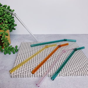 Puede incluir: Seis pajitas para beber de vidrio en varios colores, incluyendo verde, naranja, amarillo y rosa, est&aacute;n dispuestas sobre una superficie blanca con un patr&oacute;n repetido del texto "#ONEINVISIBLEEYEONDESIGN".