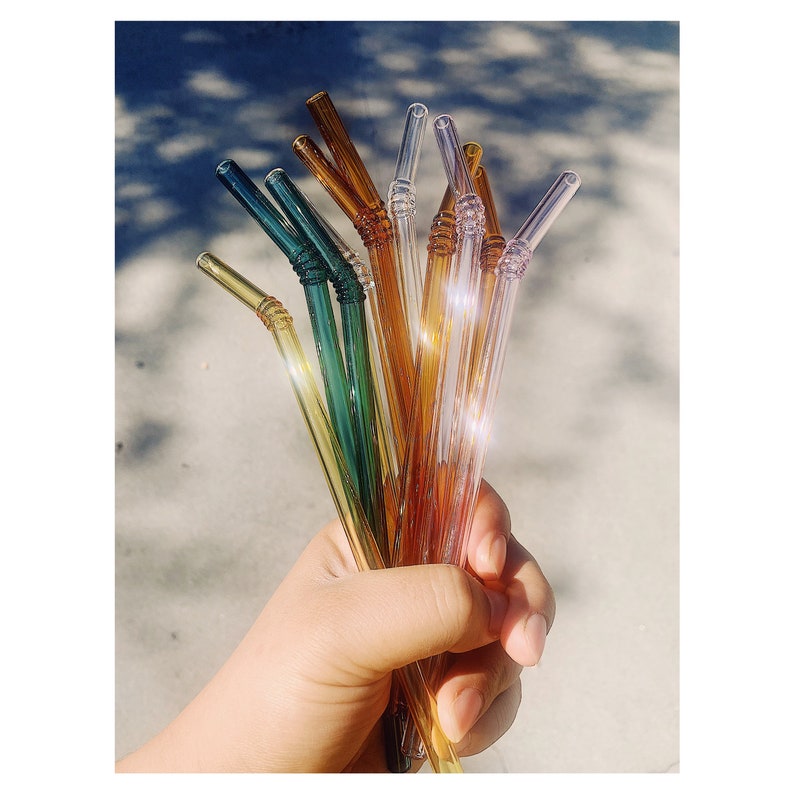 Puede incluir: Una colecci&oacute;n de pajitas para beber de vidrio de colores en varios tonos de verde, marr&oacute;n y transparente. Las pajitas est&aacute;n agrupadas y sostenidas en una mano.