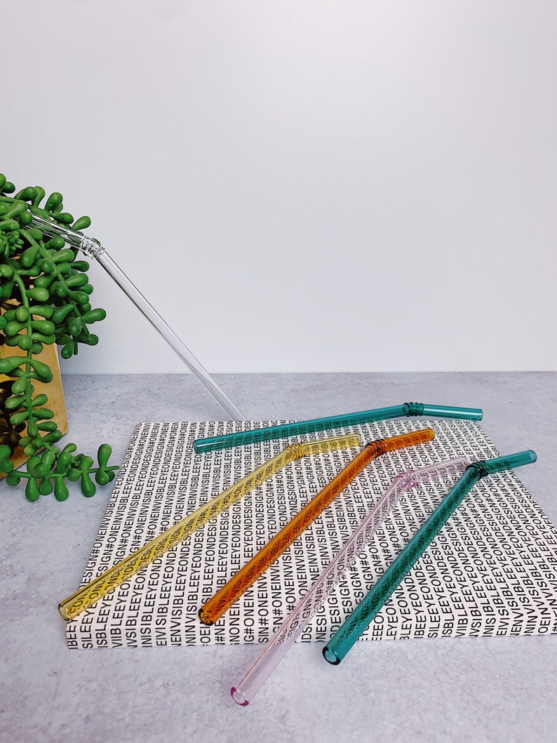 Puede incluir: Seis pajitas para beber de vidrio en varios colores, incluyendo verde, naranja, amarillo y rosa, est&aacute;n dispuestas sobre una superficie blanca con un patr&oacute;n repetido del texto "#ONEINVISIBLEEYEONDESIGN".