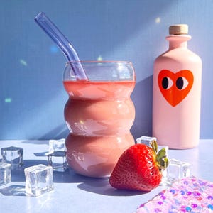 Może przedstawiać: Różowy kubek z różowym smoothie w środku, fioletową słomką ze szkła, truskawką, kostkami lodu i różową butelką z czerwonym sercem i oczami.
