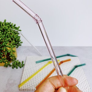 Puede incluir: Una pajita para beber de vidrio rosa con una banda texturizada cerca de la parte superior. La pajita se sostiene en una mano, y otras pajitas de colores son visibles en el fondo.