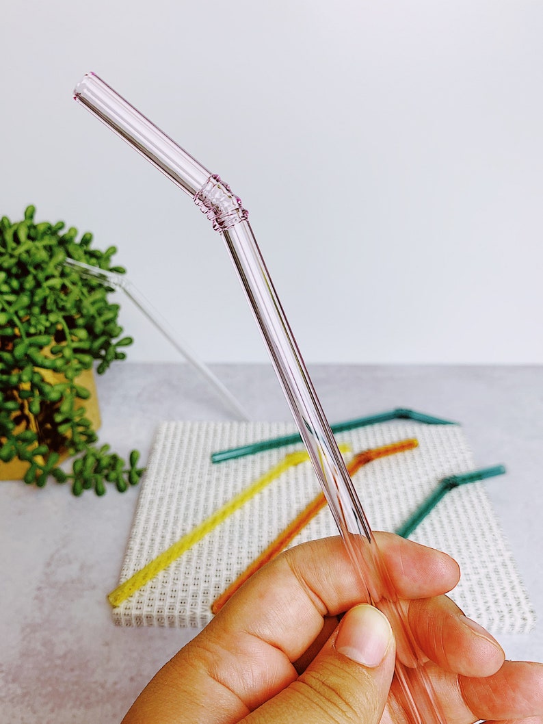 Puede incluir: Una pajita para beber de vidrio rosa con una banda texturizada cerca de la parte superior. La pajita se sostiene en una mano, y otras pajitas de colores son visibles en el fondo.