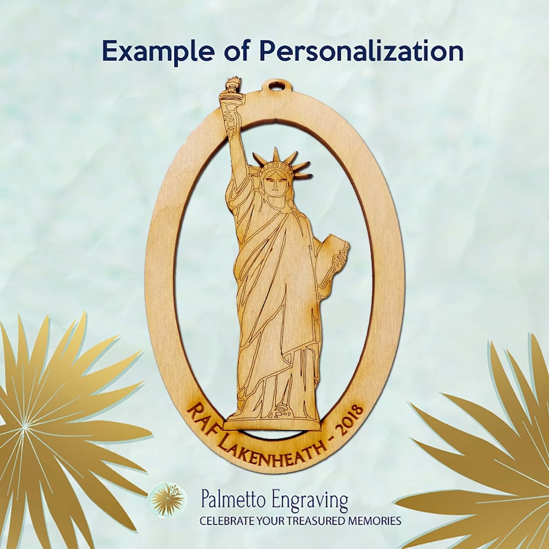 May include: A wooden oval ornament featuring a laser-cut image of the Statue of Liberty. The ornament is personalized with the text "RAF LAKENHEATH - 2018". The ornament is made by Palmetto Engraving, which is printed below the image.