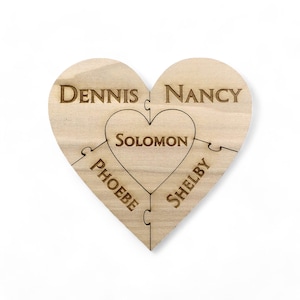 May include: A wooden heart-shaped puzzle with five interlocking pieces. Each piece has a name engraved on it: Dennis, Nancy, Solomon, Phoebe, and Shelby.