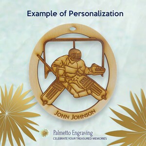 May include: A wooden hockey goalie ornament with a personalized name "John Johnson" on the bottom. The ornament features a detailed silhouette of a hockey goalie in action, with a net behind them. The ornament is a light brown color and has a rustic, handcrafted look.
