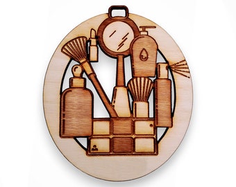 May include: A wooden ornament shaped like a circle with a cutout design of various makeup and beauty products, including a mirror, brushes, bottles, and a compact. The ornament is a light brown color.