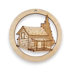 May include: A wooden ornament shaped like a circle with a cutout of a cabin and a pine tree. The ornament is made of light brown wood and has a rustic look.