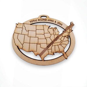 May include: A wooden ornament shaped like the United States with a dotted line across the middle showing the path of the total solar eclipse on April 8, 2024. The text "TOTAL ECLIPSE APRIL 8, 2024" is inscribed around the top of the ornament.