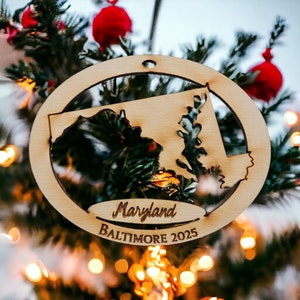 May include: A wooden Christmas ornament shaped like the state of Maryland with the words "Maryland" and "Baltimore 2025" inscribed on it. The ornament is hanging on a Christmas tree.
