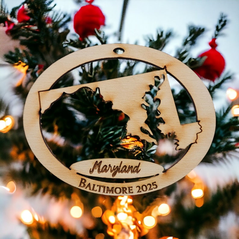 May include: A wooden Christmas ornament shaped like the state of Maryland with the words "Maryland" and "Baltimore 2025" inscribed on it. The ornament is hanging on a Christmas tree.