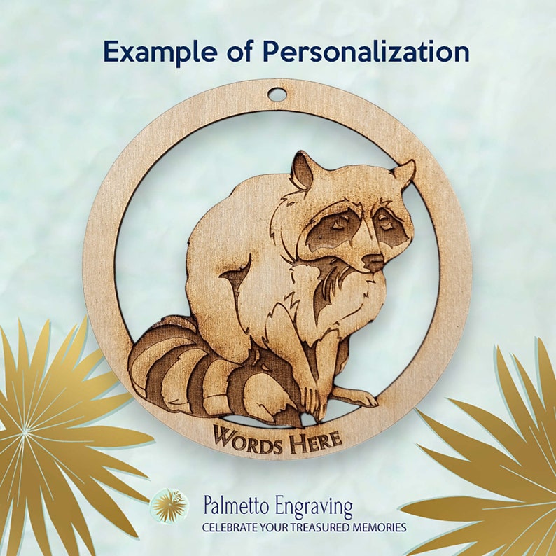 May include: A wooden ornament featuring a raccoon with a brown and black striped tail. The ornament is round and has a hole at the top for hanging. The words "WORDS HERE" are engraved below the raccoon.