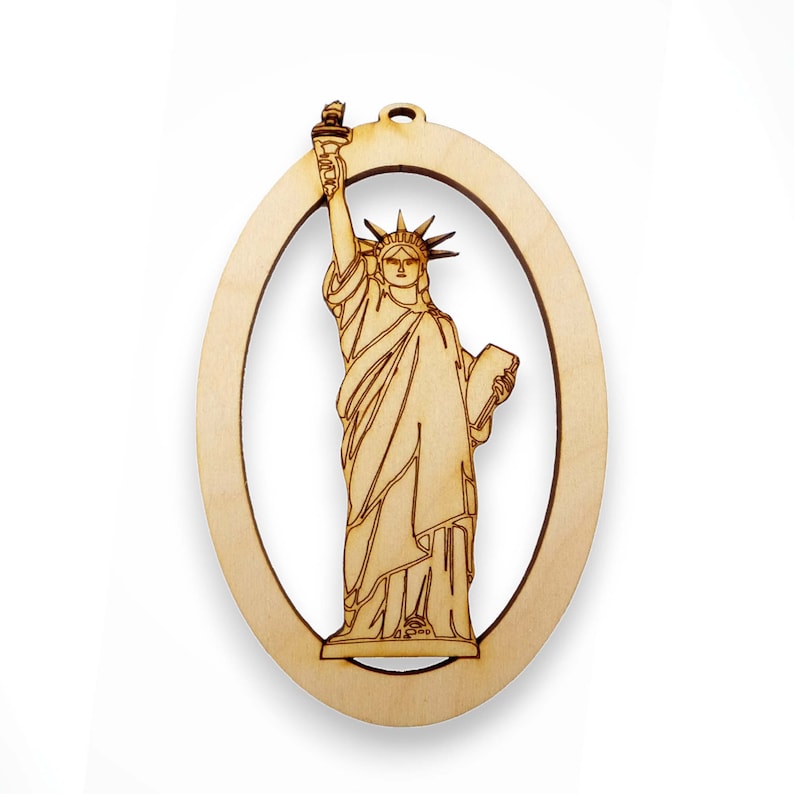 May include: A wooden ornament shaped like an oval with a cutout of the Statue of Liberty. The ornament is made of light brown wood and has a rustic look.
