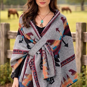 May include: A gray poncho with a Southwestern pattern featuring arrows, geometric shapes, and a tie closure. The poncho has accents of red, orange, and black. The person is wearing a brown cowboy hat and a turquoise necklace.