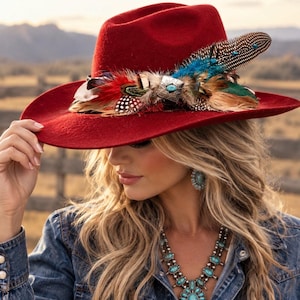 May include: A deep red felt fedora hat featuring a wide brim and a decorative band composed of assorted multi-colored feathers, including spotted and iridescent blue plumes, centered with a square silver-toned metal concho with a turquoise stone.