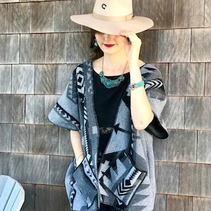 Può includere: Una donna indossa un cappello da cowboy beige con una "U" nera sul davanti, un top nero e un poncho a fantasia grigio e nero. Indossa anche gioielli turchesi, tra cui una collana e un braccialetto.