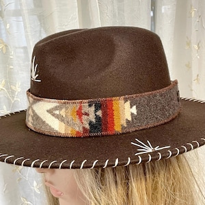 May include: A brown fedora-style hat with a decorative band featuring a colorful geometric pattern. The hat has a wide brim with white stitching and white decorative accents. The hat is a stylish accessory.