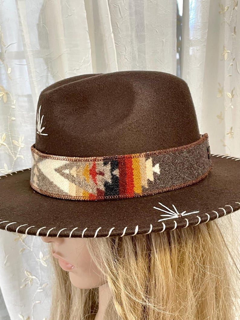 May include: A brown fedora-style hat with a decorative band featuring a colorful geometric pattern. The hat has a wide brim with white stitching and white decorative accents. The hat is a stylish accessory.