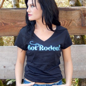 Puede incluir: Una camiseta negra con cuello en V que presenta el texto azul claro Got Rodeo? impreso en el pecho. La prenda tiene mangas cortas y un corte estándar, confeccionada en tela negra sólida con un diseño gráfico azul claro en contraste.