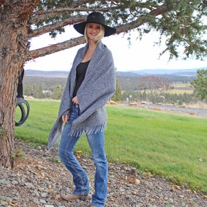May include: A woman wearing a black hat and a gray and white striped scarf stands in front of a tree. She is wearing blue jeans and brown boots. The background is a grassy field with trees in the distance.