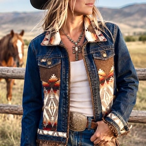 May include: A dark blue denim jacket featuring brown suede panels and a multi-colored geometric pattern on the collar, cuffs, and front. The jacket is worn over a white tank top with blue jeans, a large metal belt buckle, and a brown cowboy hat.