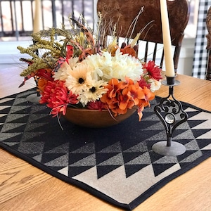 May include: A wooden table with a black and white geometric table runner. A wooden bowl filled with a fall floral arrangement is centered on the runner. A black metal candle holder with a crystal finial is on the right side of the bowl.