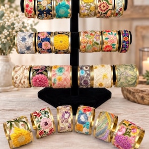 May include: A collection of cylindrical napkin rings featuring intricate floral embroidery in various colours including yellow, pink, blue, and green. Each ring has a metallic gold-toned rim and is displayed on a black T-shaped stand or arranged on a surface.