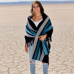 May include: A woman wearing a blue, gray, and white striped shawl with fringe. She is standing in a desert landscape and wearing blue jeans and black heeled shoes.
