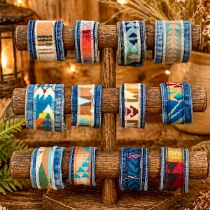 Könnte beinhalten: Ein Holzständer präsentiert neun farbenfrohe Stoffarmbänder. Jedes Armband hat ein Jeansband mit einem gemusterten Stoffmittelteil in verschiedenen Farben, darunter Blau, Orange, Rot und Türkis. Die Armbänder sind auf drei horizontalen Holzstäben angeordnet.