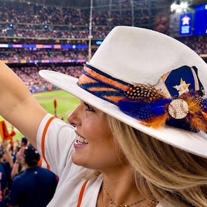 May include: A white fedora hat featuring a band with orange and navy blue tiger stripes, accented with orange and blue feathers, a Houston Astros logo patch, and a circular baseball-themed pin with a textured surface and stitching detail.
