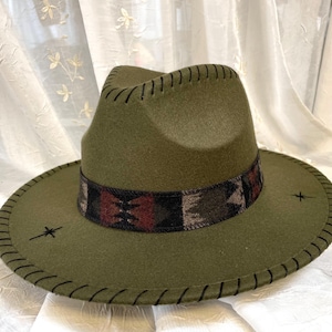May include: Olive green fedora hat with a patterned band in shades of brown, red, and black. The hat features a stitched brim and decorative stitching around the crown. Two small embroidered starburst designs are on the brim.