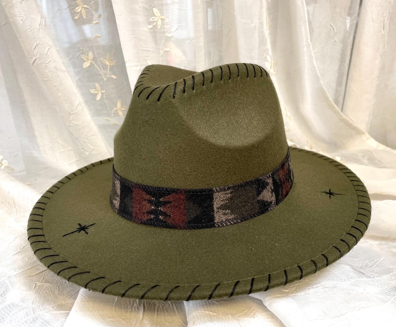 May include: Olive green fedora hat with a patterned band in shades of brown, red, and black. The hat features a stitched brim and decorative stitching around the crown. Two small embroidered starburst designs are on the brim.