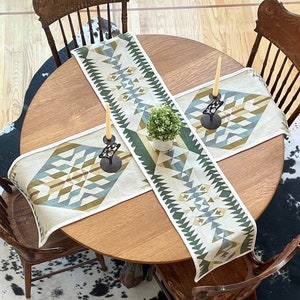 May include: A round wooden table with a black and white cowhide rug underneath. The table is set with two green and blue geometric patterned table runners and two black candle holders with white candles.